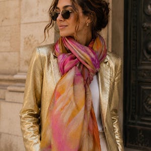 Peut inclure: Un blazer doré métallisé porté sur un haut blanc, associé à une écharpe légère avec un imprimé aquarelle abstrait dans des tons vifs de rose, d'orange et de jaune. L'écharpe présente une finition à bords effilochés et est drapée autour du cou.