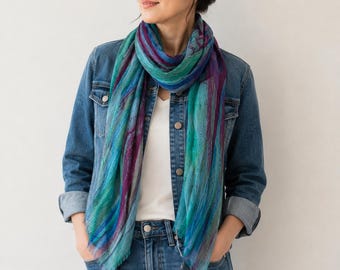 Puede incluir: Una bufanda ligera con un patrón abstracto vibrante en tonos verde azulado, turquesa, morado intenso y azul. La bufanda tiene una textura suave y semitransparente con bordes deshilachados, usada sobre una camiseta blanca y una chaqueta vaquera.