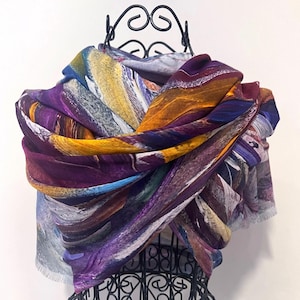 May include: A patterned scarf in shades of purple, yellow, and orange, displayed on a black metal stand. The scarf's design has a flowing, abstract quality, with visible brushstroke-like patterns. The fabric appears soft and lightweight.