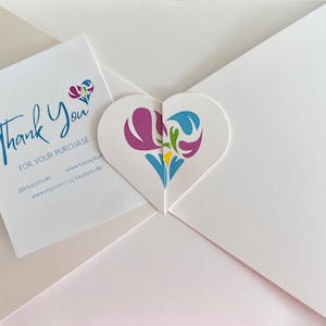 A heart-shaped custom gift envelope with a colorful floral design included with a white thank you card with blue text that reads 'Thank You for your purchase @kayjaystudio www.kayjaystudio www.etsy.com/ca/kayjaystudio'.