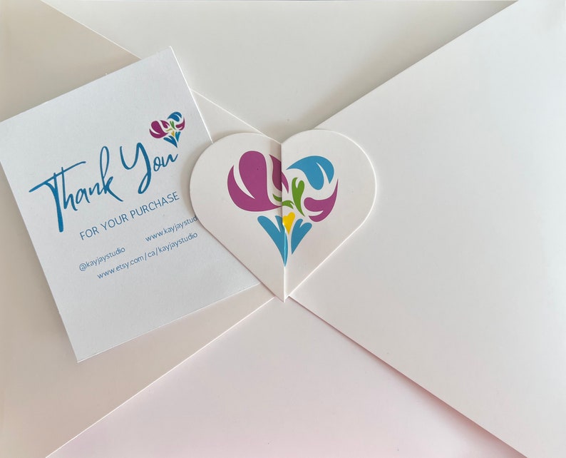A heart-shaped custom gift envelope with a colorful floral design included with a white thank you card with blue text that reads 'Thank You for your purchase @kayjaystudio www.kayjaystudio www.etsy.com/ca/kayjaystudio'.