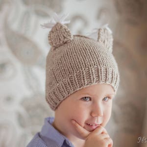 Può includere: Un cappello di lana beige con due orecchie di pelliccia bianca. Il cappello è indossato da un bambino con i capelli castani e gli occhi marroni. Il bambino indossa una camicia blu.