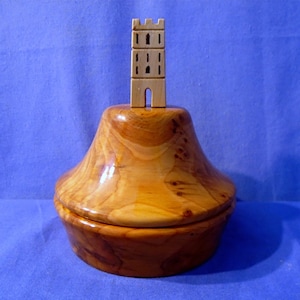 May include: A wooden box with a lid shaped like a bell. The box is made of light brown wood with a natural finish. A small wooden tower sits on top of the lid.
