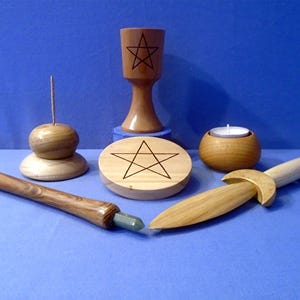 May include: A collection of wooden ritual tools, including a chalice, a round candle holder, a wooden dagger, a wooden wand with a green crystal, a wooden incense holder, and a wooden pentacle. The items are arranged on a blue surface.