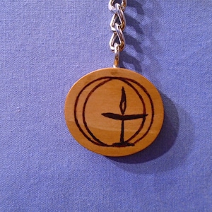 May include: A wooden pendant keychain featuring a religious symbol. The round pendant has a light brown finish and a black design of a cross with a flame above it, encircled by two rings. It is attached to a silver chain.