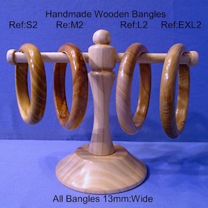 Puede incluir: Cuatro pulseras de madera hechas a mano, cada una de 13 mm de ancho, expuestas en un soporte de madera. Las pulseras varían en color, desde marrón claro hasta oscuro. El soporte es de madera clara con base redonda. El texto dice "Handmade Wooden Bangles."