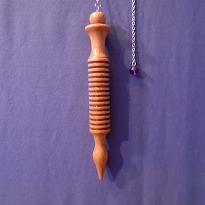 Può includere: Un pendolo in legno chiaro con scanalature orizzontali. Una catena d'argento è attaccata alla parte superiore, con una piccola perla viola all'estremità. Lo sfondo è di colore viola.