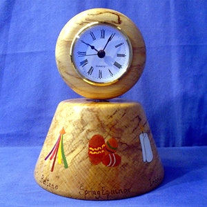 May include: A wooden clock with a round face and Roman numeral markings. The clock is mounted on a wooden base with painted images of a candle, a star, and a heart. The base is inscribed with the words "Spring Equinox", "Betane", and "Inble".