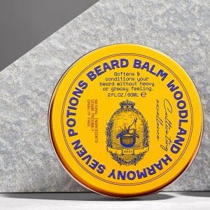 May include: A yellow tin with blue lettering that reads "Seven Potions Woodland Harmony Cultivating Excellence Beard Balm". The tin has a small illustration of a cauldron with a plant growing out of it. The text also reads "Softens & conditions your beard without heavy or greasy feeling. 2 fl oz / 60 ml e. Natural Ingredients Vegan Friendly Cruelty Free."