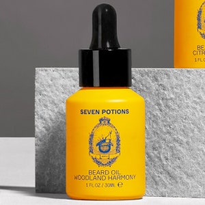 Peut inclure: Une bouteille en verre jaune avec un compte-gouttes noir et une étiquette dorée. L'étiquette indique "SEVEN POTIONS" et "BEARD OIL WOODLAND HARMONY 1 FL OZ/30ML e".