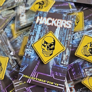 May include: A collection of collectible pins featuring a yellow skull design on a black background. The pins are square-shaped and have the word "HACKERS" printed on the backing card. The background of the card has a blue and purple digital design.