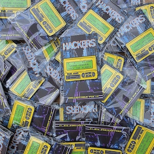 May include: A collection of "Hackers" collectible pins. Each pin features a yellow cassette tape design with the text "Grand Central Hack the Planet." The pins are packaged in clear plastic.