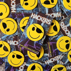 May include: A collection of yellow smiley face pins, some with an eye patch, scattered over cards with the word "HACKERS" and a circuit board design. The pins are collectible and numbered.