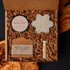 Könnte beinhalten: Eine Geschenkbox mit Schönheitsprodukten. Sie enthält ein Glas Zuckerscrub, einen schneeförmigen Gegenstand, ein Stück Seife mit Orange, Nelken und Zimt sowie einen Lippenbalsam. Die Box ist mit braunem Papier gefüllt.