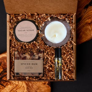 May include: A gift box filled with self-care items. Includes a sugar scrub, a candle, a spiced rum soap bar, and a rollerball bottle with flowers. The items are nestled in brown paper shreds inside a cardboard box.