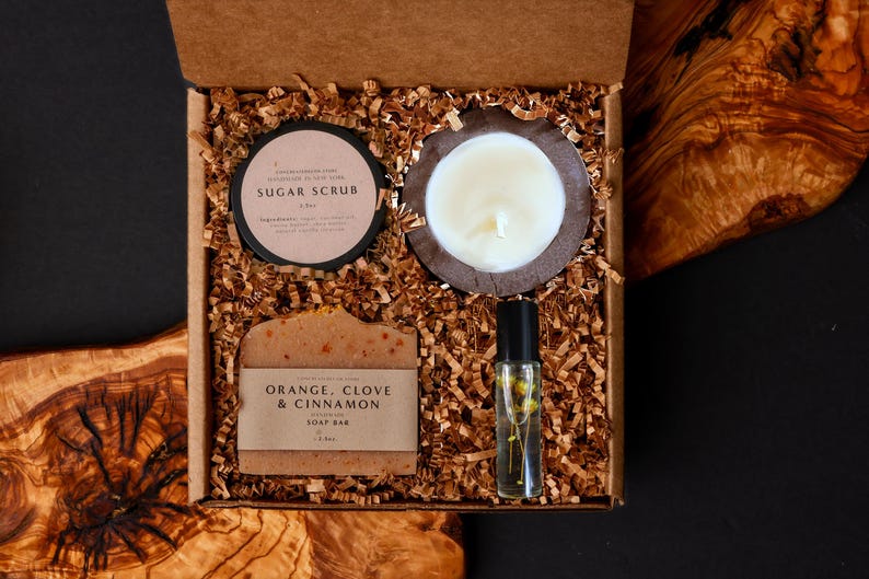 May include: A gift box filled with self-care items. The box contains a sugar scrub, a candle, a soap bar labeled "Orange, Clove & Cinnamon", and a roller bottle. The items are nestled in brown paper filler.