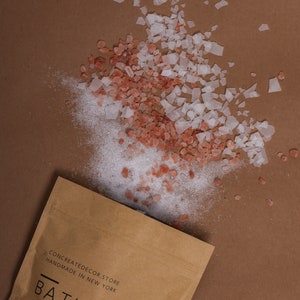 May include: A brown paper bag with the text "CONCREATEDECOR.STORE HANDMADE IN NEW YORK" and "BATH SALT" printed on it. Pink Himalayan salt and white Epsom salt are scattered on a brown surface.