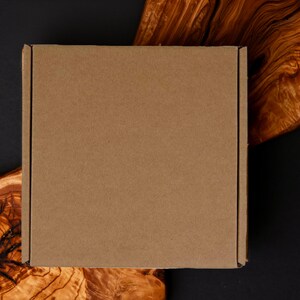 May include: A square, brown cardboard box, likely for shipping, sits on a dark surface with a natural wood grain pattern. The wood has a rich, warm color, contrasting with the box's plain surface. The background is a dark, solid color.