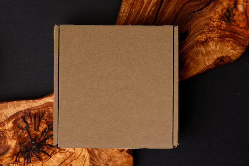 May include: A square, brown cardboard box, likely for shipping, sits on a dark surface with a natural wood grain pattern. The wood has a rich, warm color, contrasting with the box's plain surface. The background is a dark, solid color.