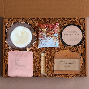 May include: A gift box containing self-care products. It features a candle, sugar scrub, clay mask, lip balm, a soap bar, and a bag of dried flowers. The items are arranged in a cardboard box with brown paper filler.
