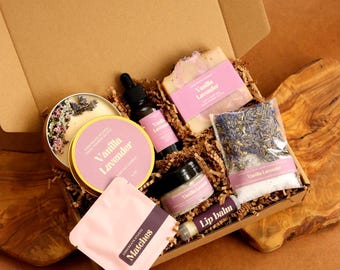 May include: A gift box containing a Vanilla Lavender candle with dried flowers, a dropper bottle, a bar of soap, a small jar, a bag of bath salts with lavender buds, a lip balm tube, and a packet of matches, all featuring pink labels.