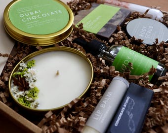 May include: A gift box containing a gold-toned tin candle with dried flowers, a Dubai Chocolate tin, a Body Scrub tin, a Body Oil glass bottle with a black dropper, a Lip Balm tube, and a rectangular dark bar, all nestled in brown paper filler.