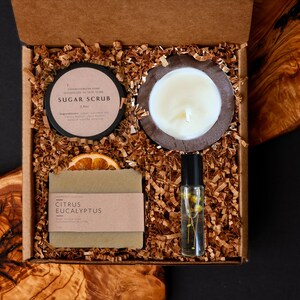 May include: A gift box filled with self-care items. Includes a sugar scrub, a candle in a wooden holder, a bar of soap with a citrus eucalyptus label, a roller bottle with flowers, and a dried orange slice. The box is filled with brown paper.