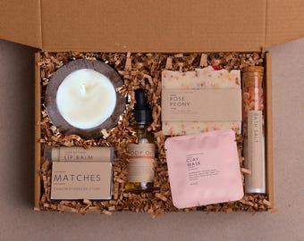 May include: A gift box filled with self-care items. Includes a candle, lip balm, matches, body oil, rose peony soap, clay mask, and bath salts. The items are nestled in brown paper and arranged in a cardboard box.