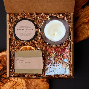 Könnte beinhalten: Geschenkbox mit Pflegeprodukten. Enthält ein Zuckerscrub, eine Kerze in einem Holzhalter, ein Seifenstück mit einem Citrus-Eukalyptus-Etikett, eine getrocknete Orangenscheibe und einen Beutel Badesalz. Die Box ist mit braunen Papierschnipseln gefüllt.