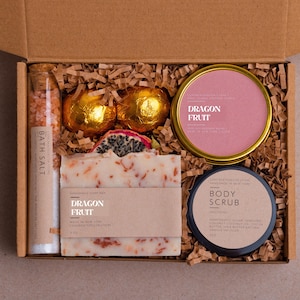 May include: A gift box containing a glass tube of bath salt, two gold-wrapped bath bombs, a pink candle tin labeled Dragon Fruit, a black jar of body scrub, and a rectangular soap bar with a label reading Dragon Fruit, all nestled in brown paper.