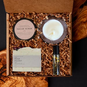 May include: A gift box filled with self-care items. The box contains a sugar scrub, a candle in a wooden holder, a bar of Rose Peony soap, and a roller bottle with flowers. The box is filled with brown paper shreds.