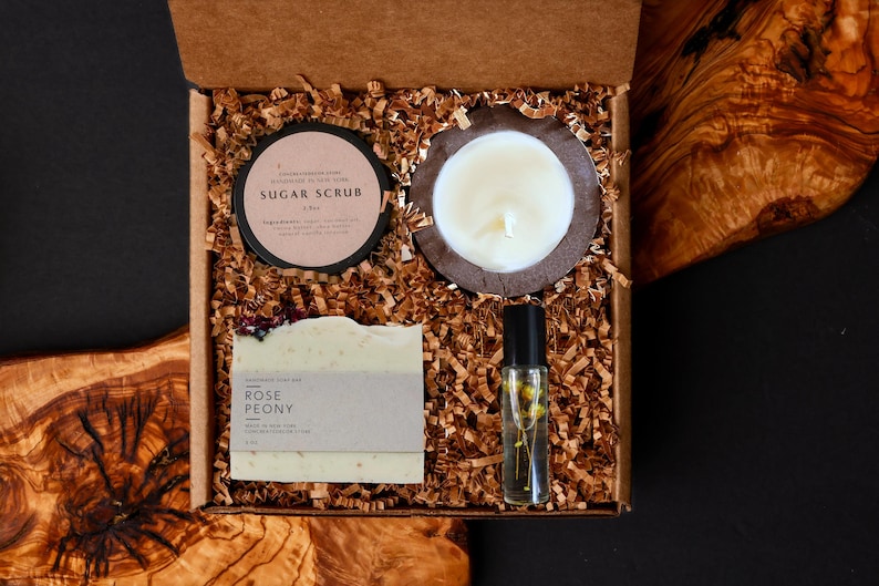 May include: A gift box filled with self-care items. The box contains a sugar scrub, a candle in a wooden holder, a bar of Rose Peony soap, and a roller bottle with flowers. The box is filled with brown paper shreds.