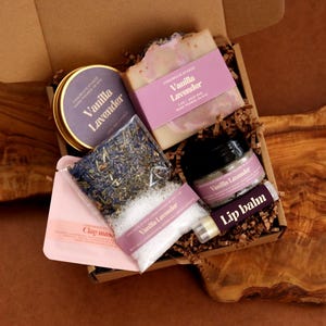 Peut inclure: Un coffret cadeau contenant des articles parfumés à la vanille et à la lavande : une bougie en boîte ronde, un savon rectangulaire, un petit pot, un baume à lèvres, un masque à l'argile et un sachet de fleurs de lavande séchées et de sels de bain blancs.