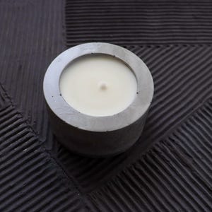 May include: A round, gray concrete candle holder with a white candle inside. The candle has a single wick. The candle holder sits on a textured, dark gray surface.