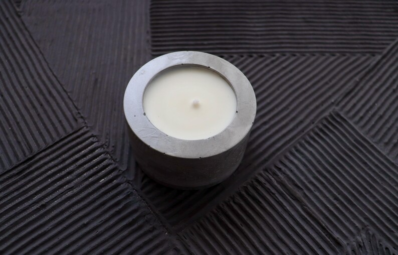 May include: A round, gray concrete candle holder with a white candle inside. The candle has a single wick. The candle holder sits on a textured, dark gray surface.