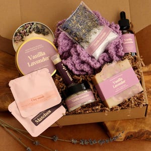 May include: A gift box containing a Vanilla Lavender candle in a gold tin, a purple crocheted washcloth, a bag of dried lavender, a Vanilla Lavender dropper bottle, a bar of soap, a lip balm, a small jar, and two pink packets labeled Clay Mask and Matches.