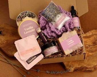 May include: A gift box containing a Vanilla Lavender candle in a gold tin, a purple crocheted washcloth, a bag of dried lavender, a Vanilla Lavender dropper bottle, a bar of soap, a lip balm, a small jar, and two pink packets labeled Clay Mask and Matches.