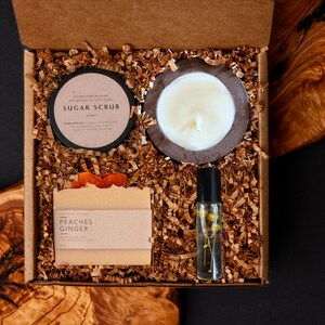 May include: A gift box filled with self-care items. Includes a sugar scrub, a candle in a wooden holder, a bar of soap labeled "Peaches Ginger", and a roller bottle with flowers. The box is filled with brown paper shreds.