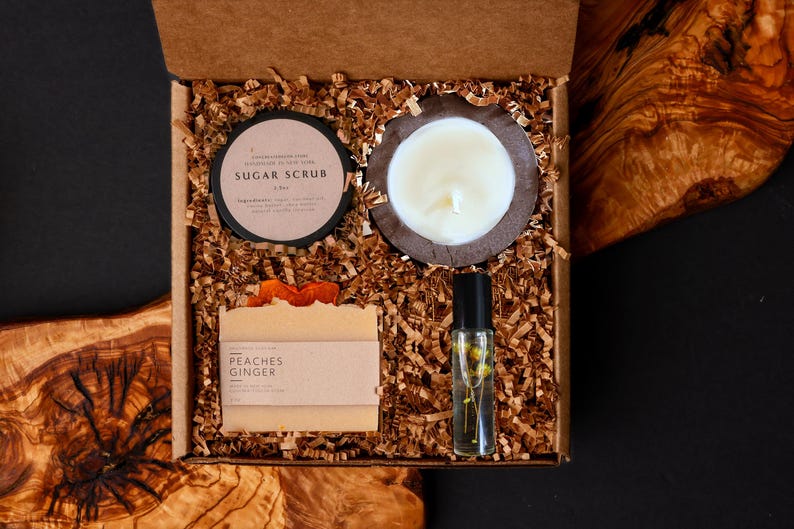 May include: A gift box filled with self-care items. Includes a sugar scrub, a candle in a wooden holder, a bar of soap labeled "Peaches Ginger", and a roller bottle with flowers. The box is filled with brown paper shreds.