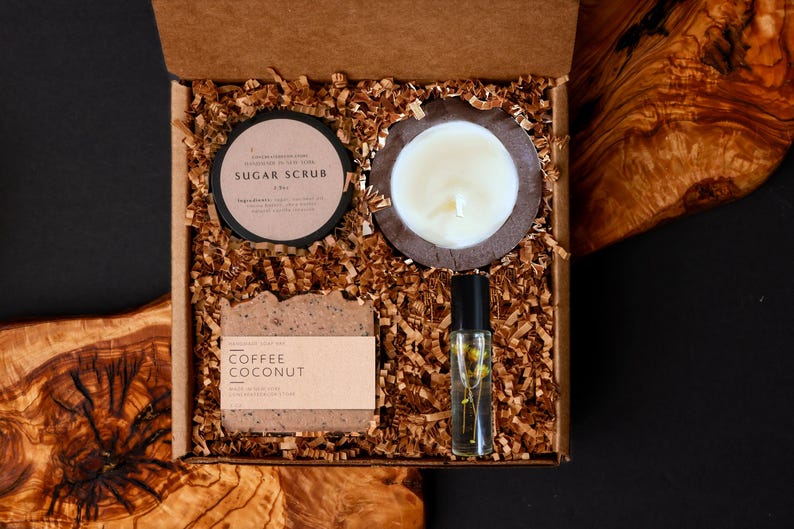 May include: A gift box filled with self-care items. Includes a sugar scrub, a candle, a bar of coffee coconut soap, and a roller bottle with a floral design. The items are nestled in brown paper shreds inside a cardboard box.