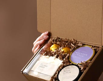 May include: A brown cardboard gift box filled with brown paper crinkle filler containing a bar of Leather Amber soap, a round black container labeled Body Scrub, a round gold tin labeled Lavender Vanilla, and two gold foil-wrapped spherical items.