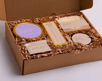 May include: A brown cardboard box filled with a lavender vanilla bath melt, a lavender vanilla soap bar, a shea butter jar, a lip balm, and a box of matches. The box is filled with brown shredded paper.