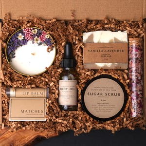 May include: A gift box containing a candle topped with dried flowers, a bottle of body oil, a bar of vanilla-lavender soap, a tube of lip balm, a box of matches, a jar of sugar scrub, and a glass vial filled with pink and white bath salts.