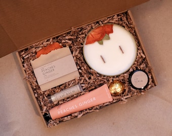 May include: A gift box filled with self-care items, including a candle with dried flower petals, a bar of soap, a tube of hand cream, a lip balm, and a lip scrub. The box is filled with brown paper shreds and the items are labeled "Peaches Ginger".