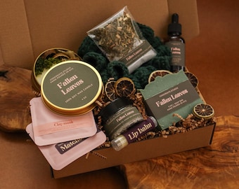 May include: A gift box containing a Fallen Leaves soy wax candle in a gold tin, a Fallen Leaves soap bar, a small jar, a lip balm, two pink clay mask packets, a dark green knitted item, a dropper bottle, and dried citrus slices on brown paper filler.