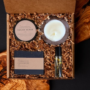 May include: A gift box filled with self-care items. Includes a sugar scrub, a candle in a coconut shell, a bar of soap labeled "Tobacco Cashmere", and a roller bottle. The box is filled with brown paper shreds.