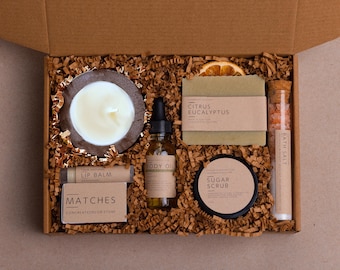 May include: A gift box filled with self-care items. Includes a candle, lip balm, body oil, soap, sugar scrub, bath salts, and matches. The items are arranged on shredded paper inside a cardboard box.