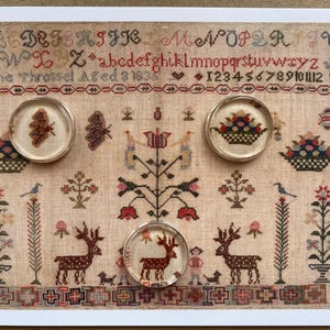 May include: A rectangular sampler featuring cross-stitch embroidery on beige fabric, displaying the alphabet, numbers, and the text Jane Throssel Aged 9 1838. Three circular glass paperweights are placed on top, showing a butterfly, a basket, and a deer.