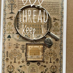 Puede incluir: Una pulsera plateada con un dije de corazón y un pequeño colgante cuadrado con un diseño de casa. La pulsera está colocada sobre un fondo de un textil bordado de estilo vintage con texto.