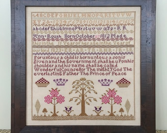 May include: A cross-stitch sampler with a beige background and brown, gold, and purple embroidery. The sampler features a verse from the Bible, "For unto us a child is born, unto us a son is given: and the government shall be upon his shoulder: and his name shall be called Wonderful, Counsellor, The mighty God, The everlasting Father, The Prince of Peace."